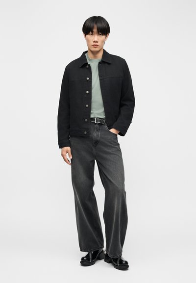 Νεαρό άτομο που φοράει μαύρο σακάκι με κουμπιά, πράσινο μπλουζάκι, φαρδιά γκρι τζιν, μαύρη ζώνη και λαμπερά μαύρα παπούτσια, στέκεται με το ένα χέρι στην τσέπη.