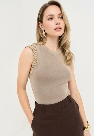 Kvinde med langt blondt hår har en ærmeløs beige top, mørkebrune bukser med høj talje, søstjerneformede øreringe og et gyldent armbånd på, poserer i profil.