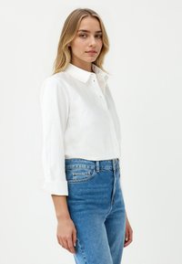 White button-up shirt with a collar, long sleeves rolled at the cuffs, paired with high-waisted blue jeans. Smooth fabric, classic design.