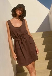 Braunes Leinenkleid mit Rundhalsausschnitt, fünf Knöpfen, einem verstellbaren Kordelzugbund und zwei aufgesetzten Taschen. Kombiniert mit einem schwarzen gehäkelten Hut.