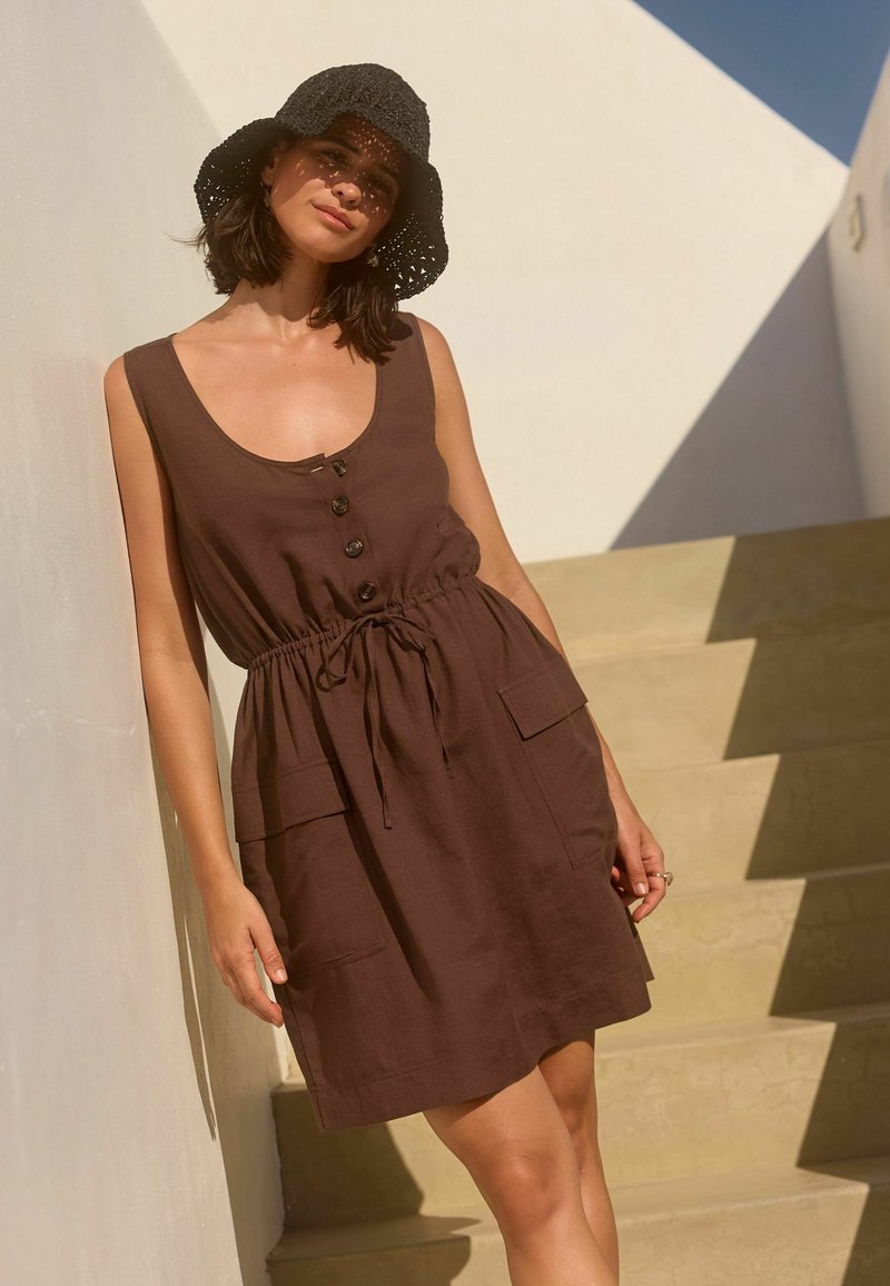 Braunes Leinenkleid mit Rundhalsausschnitt, fünf Knöpfen, einem verstellbaren Kordelzugbund und zwei aufgesetzten Taschen. Kombiniert mit einem schwarzen gehäkelten Hut.