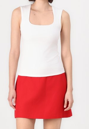 Femme portant un haut blanc sans manches avec un col carré et une jupe rouge trapèze, debout devant un fond uni.