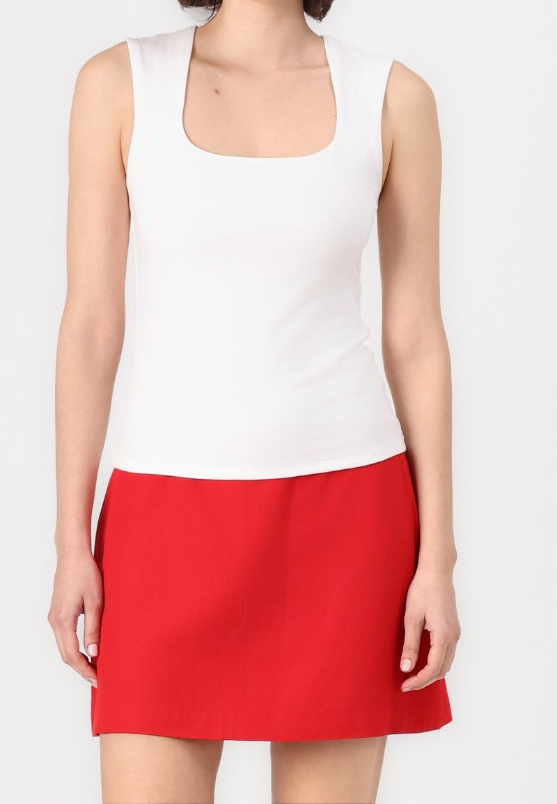 Femme portant un haut blanc sans manches avec un col carré et une jupe rouge trapèze, debout devant un fond uni.