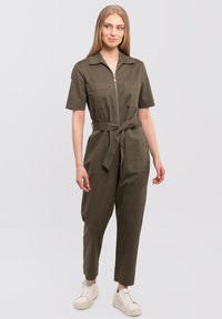 Olive green jumpsuit with short sleeves, zip front, two chest pockets, and a tie belt; paired with white sneakers. Soft, cotton blend fabric.