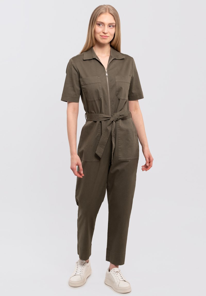 Olive green jumpsuit with short sleeves, zip front, two chest pockets, and a tie belt; paired with white sneakers. Soft, cotton blend fabric.