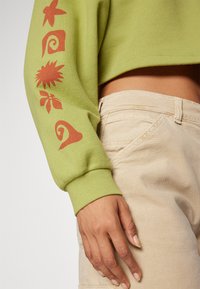 Green cropped sweatshirt with printed red patterns on the sleeve. Beige pants with a textured finish and side pockets.
