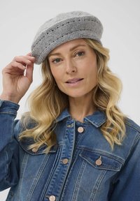 Beret gris de punto con un diseño texturizado, que presenta una banda y pequeños agujeros a lo largo de la parte superior. Combinado con una chaqueta de mezclilla azul.