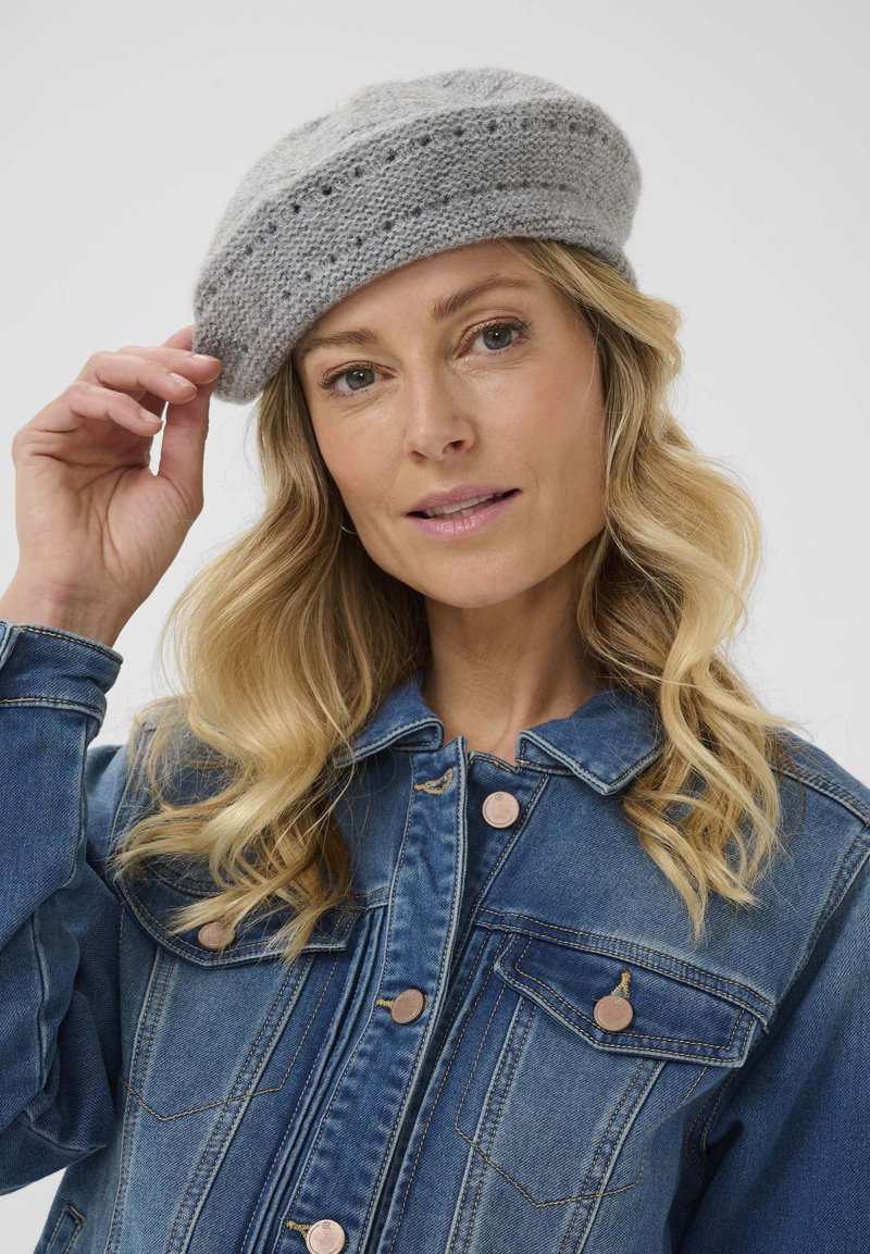 Beret gris de punto con un diseño texturizado, que presenta una banda y pequeños agujeros a lo largo de la parte superior. Combinado con una chaqueta de mezclilla azul.