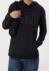 Felpa con cappuccio grigio scuro con logo "GAP" nero, dotata di cappuccio con coulisse e tasca a marsupio, abbinata a jeans di jeans e una piccola borsa nera.