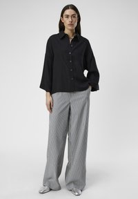 Camisa negra de manga larga con botones y corte relajado, combinada con pantalones de rayas anchas en gris y blanco, y zapatos planos plateados.