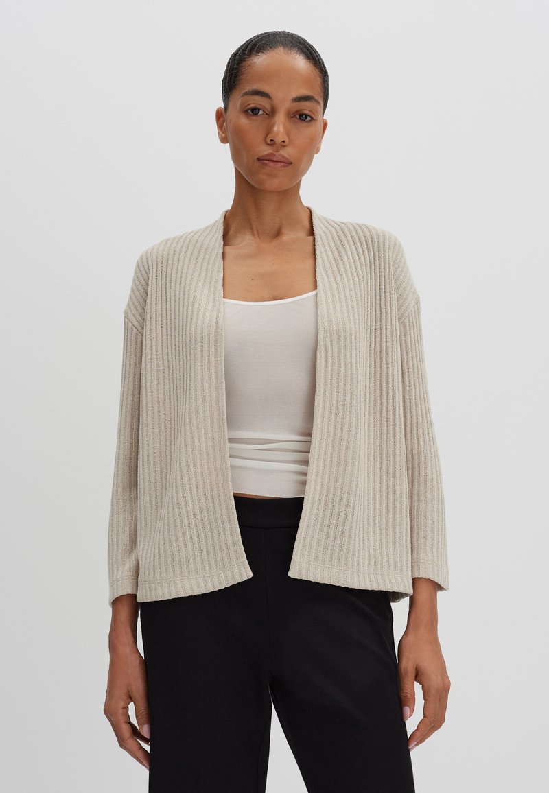 Beige geribbelde cardigan met een open voorkant, driekwartmouwen en een gestructureerde afwerking, gecombineerd met een aansluitend wit topje en zwarte broeken.