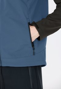 Wasserabweisende blaue Jacke mit einer Reißverschlusstasche auf der linken Seite. Die Bündchen sind schwarz mit Klettverschluss-Einstellungen. Glatte Textur.