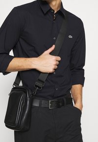 Černá košile na knoflíky s dlouhými rukávy, doplněná černou koženou crossbody taškou s lesklým povrchem, vybavenou zipem a nastavitelným popruhem.