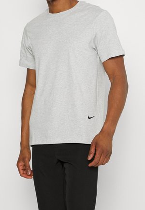 Mann trägt ein hellgraues Nike-T-Shirt mit kurzen Ärmeln und schwarze Hosen, steht vor einem schlichten weißen Hintergrund.