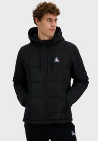 Veste en duvet noire matelassée avec capuche, dotée d'une fermeture éclair sur le devant, de poches latérales et d'un logo sur la poitrine gauche. Texture douce.