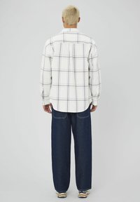 Chemise en flanelle blanche avec un motif de tartan violet, associée à un jean bleu ample et des baskets décontractées. La chemise a des manches longues et un ourlet arrondi.
