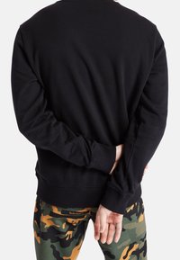 Sudadera de algodón negra con puños y dobladillo acanalados, llevada sobre pantalones de camuflaje. La persona tiene las manos entrelazadas detrás de la espalda.