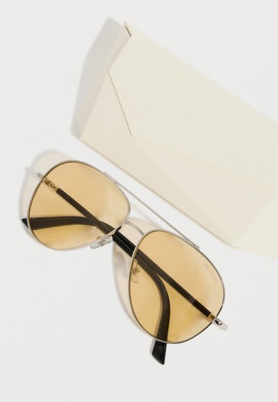 Γυαλιά ηλίου σε στιλ πιλότου με φακούς χρώματος κίτρινο και μαύρα πλαϊνά, δίπλα σε θήκη Jimmy Choo σε κρεμ χρώμα, πάνω σε λευκό φόντο.