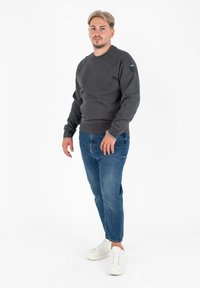 Szary sweatshirt z prążkowanymi mankietami i dołem, noszony na niebieskich dżinsach. Białe sneakersy dopełniają outfit, na jednolitym tle.