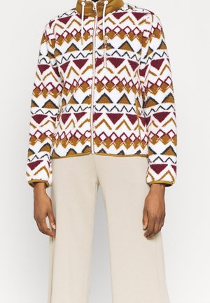 Veste polaire avec des triangles géométriques blancs, bordeaux, moutarde et noirs, fermeture éclair devant et poche poitrine, portée avec un pantalon crème.