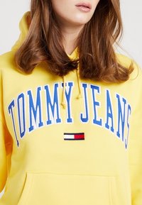 Felpa gialla con cappuccio che presenta la scritta "TOMMY JEANS" in lettere blu e un piccolo logo rosso, bianco e blu come dettaglio. Realizzata in tessuto morbido, con tasca a marsupio.