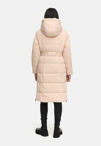 Long, blush pink puffer coat with a hood. Features quilted texture, a belted waist, and zippered side vents. Worn over dark pants and boots.