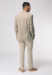 Hellbeiger zweiteiliger Anzug mit strukturiertem Finish. Verfügt über ein einreihiges Jacket, schlanke Hose und hellbraune Slippers.