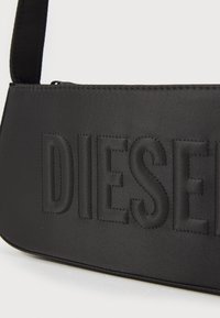 Mustat olkalaukku, joka on valmistettu kestävästä nailonista, ja jossa on kohokuvioitu "DIESEL"-logo edessä sekä vetoketjukiinnitys ylhäällä.