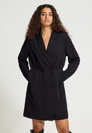 Femme aux cheveux bouclés portant une longue robe blazer noire à double boutonnage, les mains dans les poches, sur un fond uni.