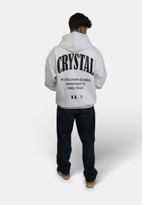 Felpa bianca con grande testo nero "CRYSTAL" sulla schiena, che presenta dettagli dell'indirizzo di Parigi. Abbinata a jeans scuri e scarpe gialle.