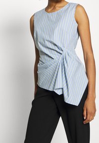 Ljusblå ärmlös topp med gula vertikala ränder. Har en draperad front med en tvistdetalj, tillsammans med svarta byxor. Glatt tyg.