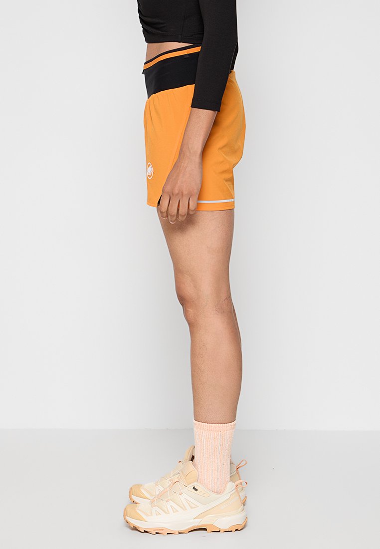 Shorts deportivos naranjas con cinturilla negra y acentos blancos, con un logo en el lateral. Combinados con zapatos beiges y calcetines rosa claro.