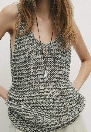 Vrouw draagt een mouwloze, losgebreide grijze top en een lange ketting met een druppelvormige hanger, handen rustend op de heupen.