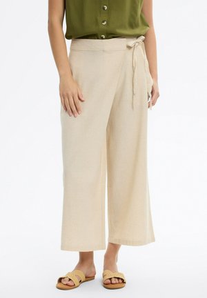 Mujer con pantalones anchos beige con cintura atada y sandalias planas tejidas, combinados con un top verde sin mangas con botones.