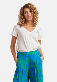 Witte katoenen V-hals t-shirt met korte mouwen, voorzien van een bloemenborduursel op de linkerkant van de borst; gecombineerd met felblauwe broeken met groene abstracte patronen.