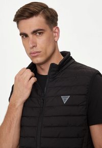 Gilet trapuntato nero con collo alto, chiusura a zip e un logo triangolare grigio sul lato sinistro del petto. Indossato sopra una maglietta nera.