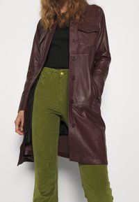 Manteau en cuir bourgogne avec un col et une fermeture à boutons, présentant une poche poitrine et des manches longues. Associé à un pantalon en velours côtelé vert.