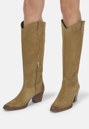 Beige Wildleder kniehohe Stiefel mit seitlichem Reißverschluss und gestapelten Holzabsätzen, getragen von einer Person vor einem schlichten hellen Hintergrund.