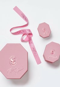 Cajas octagonales rosas con logotipos en relieve, acompañadas de una cinta rosa que muestra "SWAROVSKI" y "Ariana Grande" en letras plateadas.