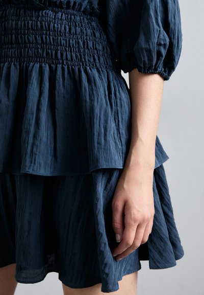 Tummansininen mekko, jossa on tekstuuri, smockattu bodice, kerrokselliset rypytykset ja puffimaiset hihat. Mekossa on valuva hame ja elegantti muotoilu.