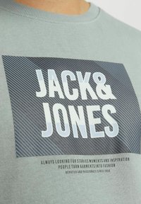 Camiseta de algodón verde clara con un estampado gráfico que presenta "JACK & JONES" en letras blancas en negrita y patrones de líneas diagonales en el fondo.