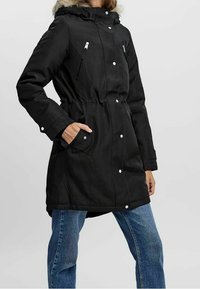 Parka noire avec une taille cintrée, capuche en fausse fourrure amovible, fermeture éclair et boutons-pression à l'avant, et deux poches latérales, associée à un jean bleu.