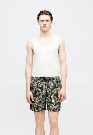 Νέος άνδρας που στέκεται προς τα εμπρός, φοράει λευκό φανελάκι και μαύρο μαγιό με πράσινο μοτίβο φύλλων φοίνικα.