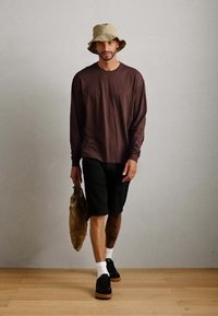 Brown long-sleeve shirt, black shorts, black sneakers with tan accents, holding a tan bag, wearing a beige bucket hat. Light wooden floor.