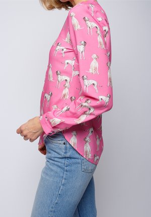 Vrouw draagt een roze blouse met lange mouwen met dalmatiër hond prints, gecombineerd met een blauwe spijkerbroek, zijaanzicht tegen een effen achtergrond.