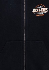 Tmavomodrá zipsová mikina s dvoma prednými vreckami, kovovým zipsom a logom "Jack & Jones" v bielej a oranžovej farbe na hrudi. Mäkká textúra látky.