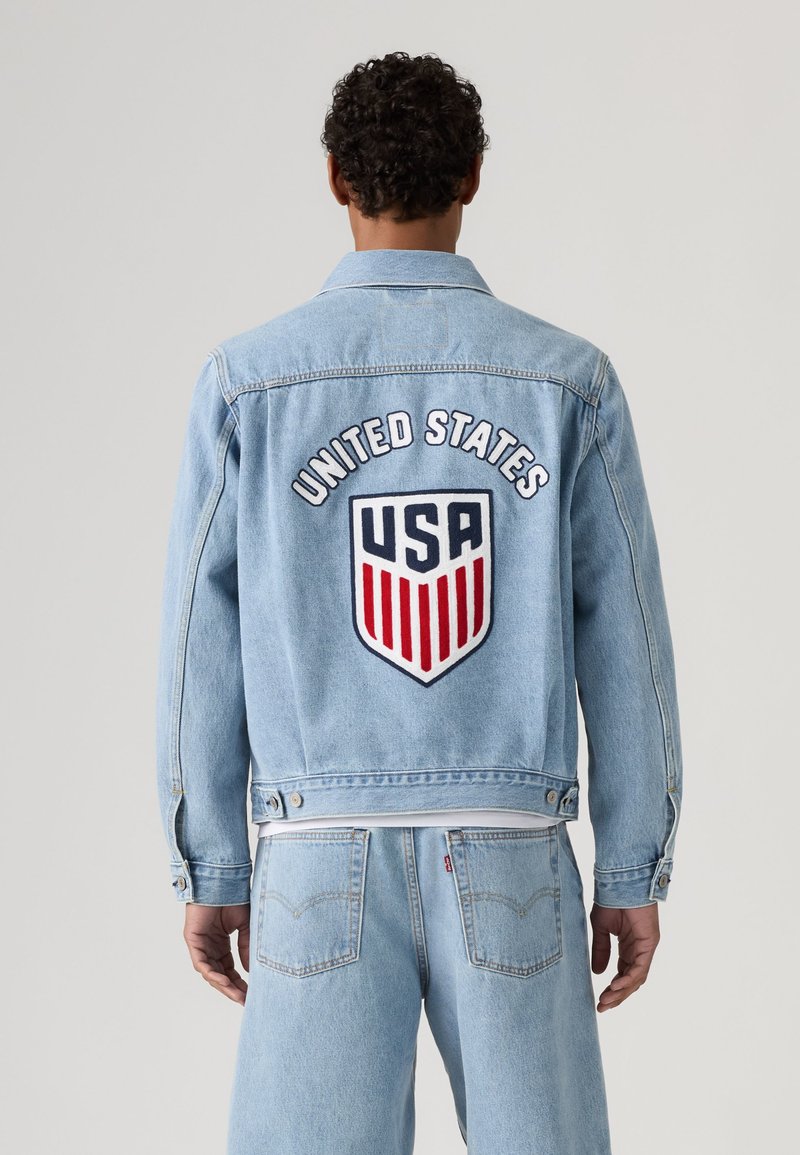 Homme portant une veste en denim bleu clair avec "United States" et un écusson bouclier des États-Unis dans le dos, assortie à un jean en denim.