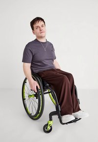 Black wheelchair with green accents, person in gray t-shirt and brown pants, seated. Smooth floor backdrop, minimalistic setting.