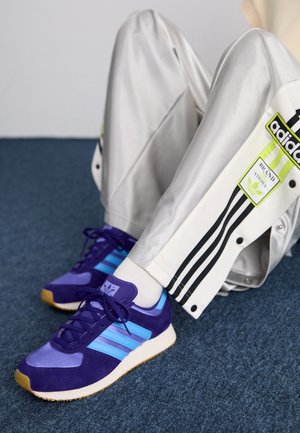 Personne portant un pantalon de survêtement Adidas argenté et des baskets Adidas violettes, assise sur un sol recouvert de moquette bleue, jambes pliées.