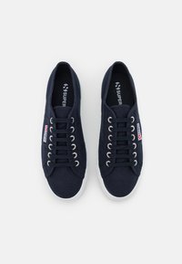 Sneakers in tela blu con suole in gomma bianca, dotati di occhielli e lacci in metallo, e un piccolo tag logo rosso e bianco sul lato.